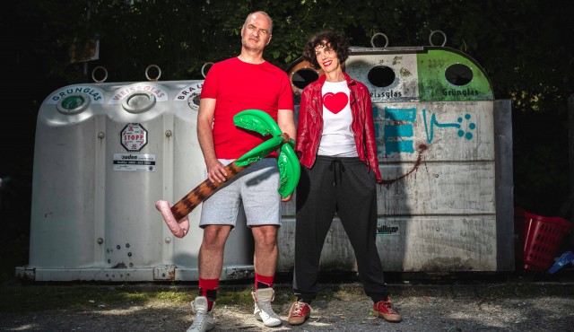 Birgit und Wolfgang mit aufblasbarer Palme in der Hand vor Altglascontainer.