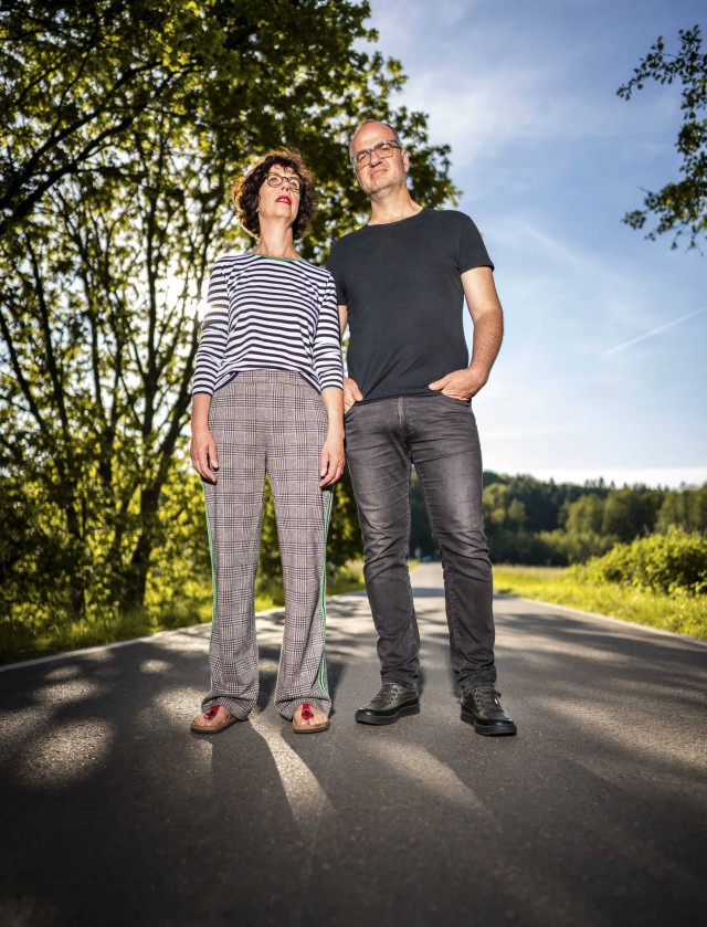 Kamera aus Froschperspektive zeigt Birgit und Wolfgang auf Landstraße stehend im Sonnenschein.