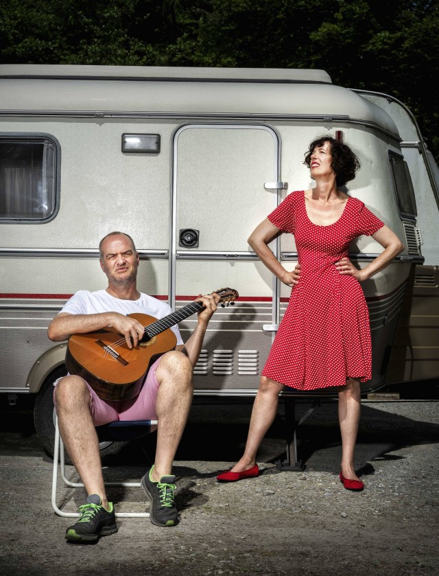 Wolfgang mit Klampfe auf Campingstuhl. Birgit in rotem Kleid und Hände auf den Hüften steht daneben. Im Hintergrund ein Wohnwagen.